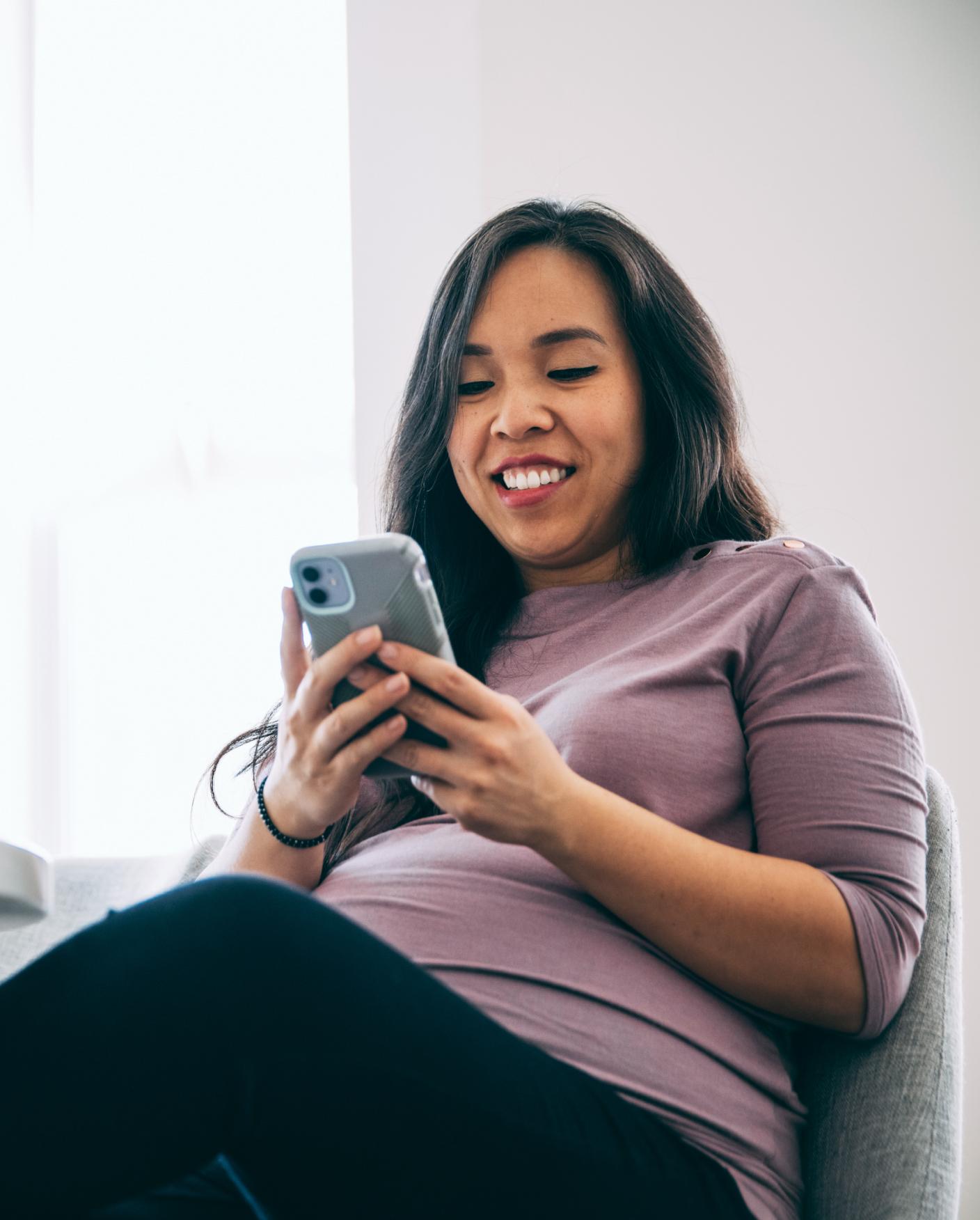 pregnant woman sits in chair and looks at phone while smiling